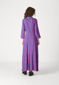 Robe longue et fluide avec un col, présentant des rayures verticales en rose et bleu. Manches longues et taille cintrée. Chaussures à talons blocs noires.