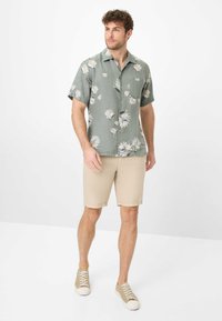 Camicetta verde a maniche corte con motivo floreale, pantaloni corti beige testurizzati e sneakers chiare. La camicia ha una tasca frontale e grandi motivi di fiori bianchi.