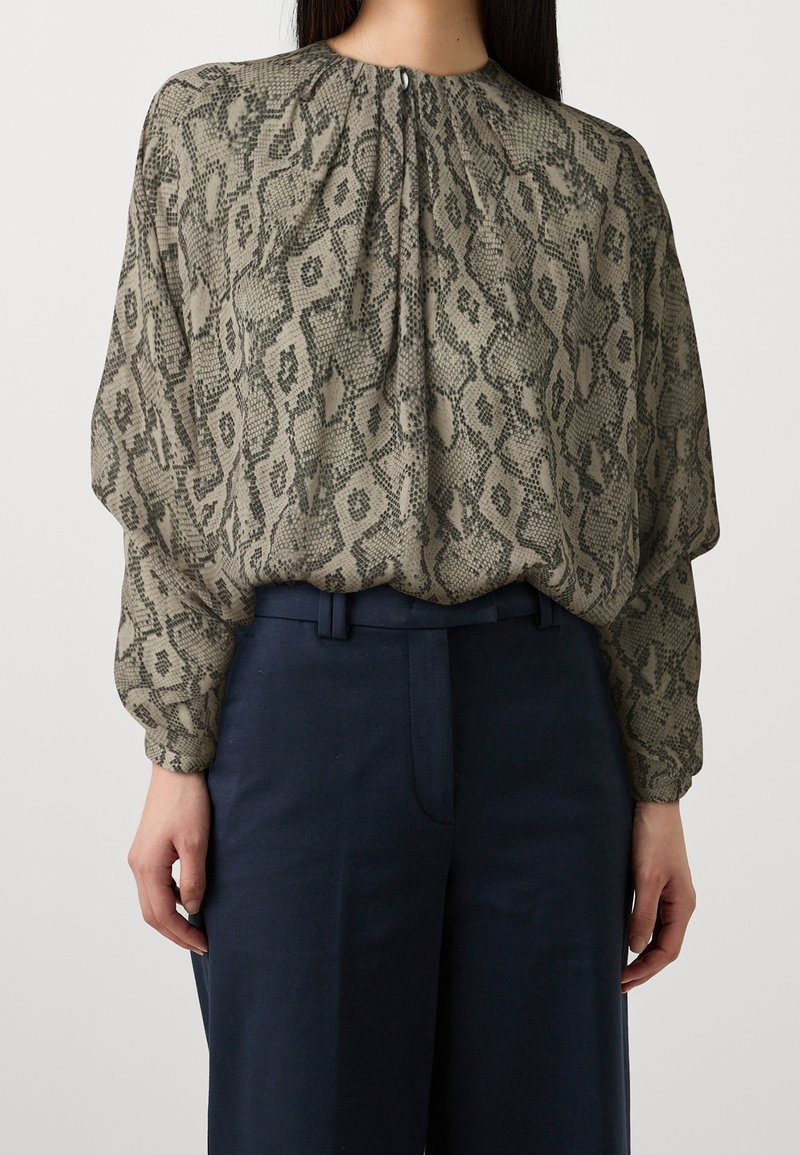 Femme portant un chemisier ample à manches longues imprimé serpent, rentré dans un pantalon taille haute bleu marine foncé, sur un fond clair uni.