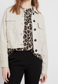 Beżowa kurtka typu cropped z dwoma kieszeniami na piersi i czarnymi guzikami, zestawiona z bluzką w panterkę. Całość dopełniają czarne spodnie.