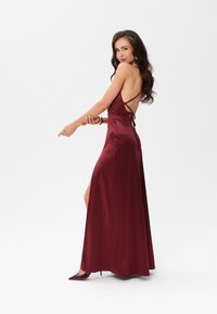 Burgundská saténová maxi šaty s odhaleným zadním dílem, tenkými ramínky a vysokým rozparkem. Modelka pózuje bokem, má na sobě odpovídající lodičky.