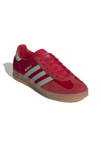 Červené semišové tenisky s kulatou špičkou, stříbrnými třemi pruhy a texturovanými akcenty na jazyku. Průhledná gumová podrážka. Viditelné logo "Gazelle".