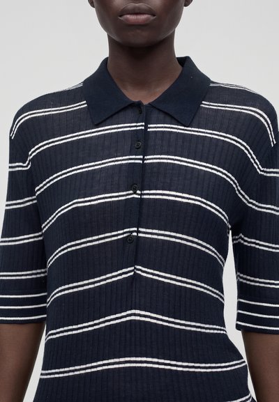 Personne portant un polo bleu marine côtelé avec des rayures blanches horizontales et des boutons noirs, montré des épaules jusqu'au milieu du torse.