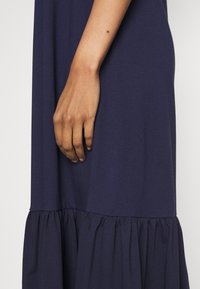 Mörkblå klänning i mjukt tyg med volangkant och avslappnad passform. En hand vilar försiktigt på ena sidan av klänningen.