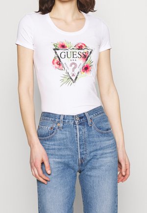 Frau trägt ein weißes, eng anliegendes T-Shirt mit farbenfrohem, floralem Guess-Logo und hoch taillierte blaue Jeans vor einfarbigem Hintergrund.