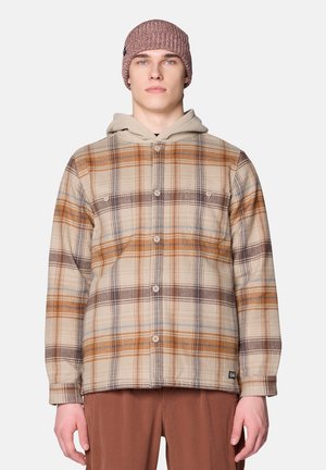 Ein junger Mann trägt eine beige-braune karierte Hemdjacke mit einem beigen Kapuzenpullover darunter und eine mauve gestrickte Mütze.