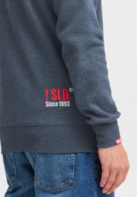 Dunkelgrauer Sweatshirt mit gerippten Bündchen und Saum. Verfügt über das rote gestickte Logo "! SLD Seit 1993" auf der unteren linken Seite. Denim-Jeans sind sichtbar.