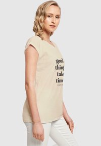 Beige puuvillane T-särk lühikeste varrukatega ja V-kaelusega, mille esiküljel on must trükitud slogan "head asjad võtavad aega".