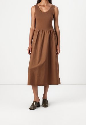 Bruine mouwloze jurk met een getailleerd lijfje en een geplooide rok; gemaakt van zachte stof. Gecombineerd met donkerglanzende hoge muiltjes.