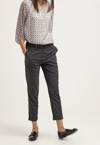 Grau tapered Hosen mit einer subtilen Textur, kombiniert mit einer durchsichtigen, gemusterten Bluse mit geometrischen Designs in Weiß und Grau. Schwarze Loafers.