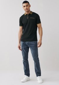 Juoda polo marškinėliai su tekstūruota apdaila, geltonos detalės ant apykaklės ir krūtinės kišenės, derinami su mėlynais džinsais ir baltais sportbačiais.