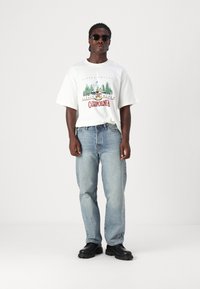 White graphic t-shirt with green and red print; light blue jeans; black sunglasses; black chunky shoes. Casual outfit, relaxed fit.