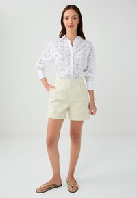Chemise blanche brodée avec découpes florales et col, assortie à un short crème mi-long avec ourlets retroussés.