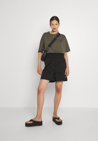 T-shirt oversize verde oliva, pantaloni corti neri con vita elastica e infradito nere con plateau. Design semplice con accenti minimalisti, texture liscia.