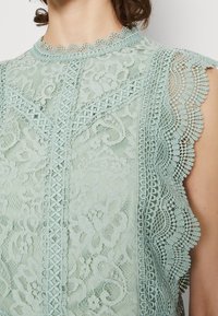 Haut en dentelle vert menthe présentant des motifs floraux complexes, des bords festonnés et des détails le long de l'encolure et des manches.