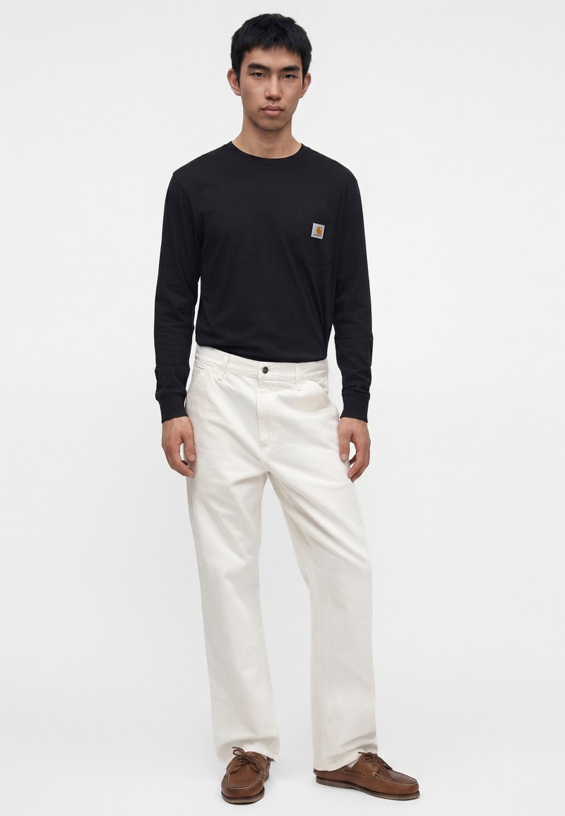 Homme portant une chemise noire à manches longues avec patch logo, pantalon blanc droit et chaussures en cuir marron, debout devant un fond uni.