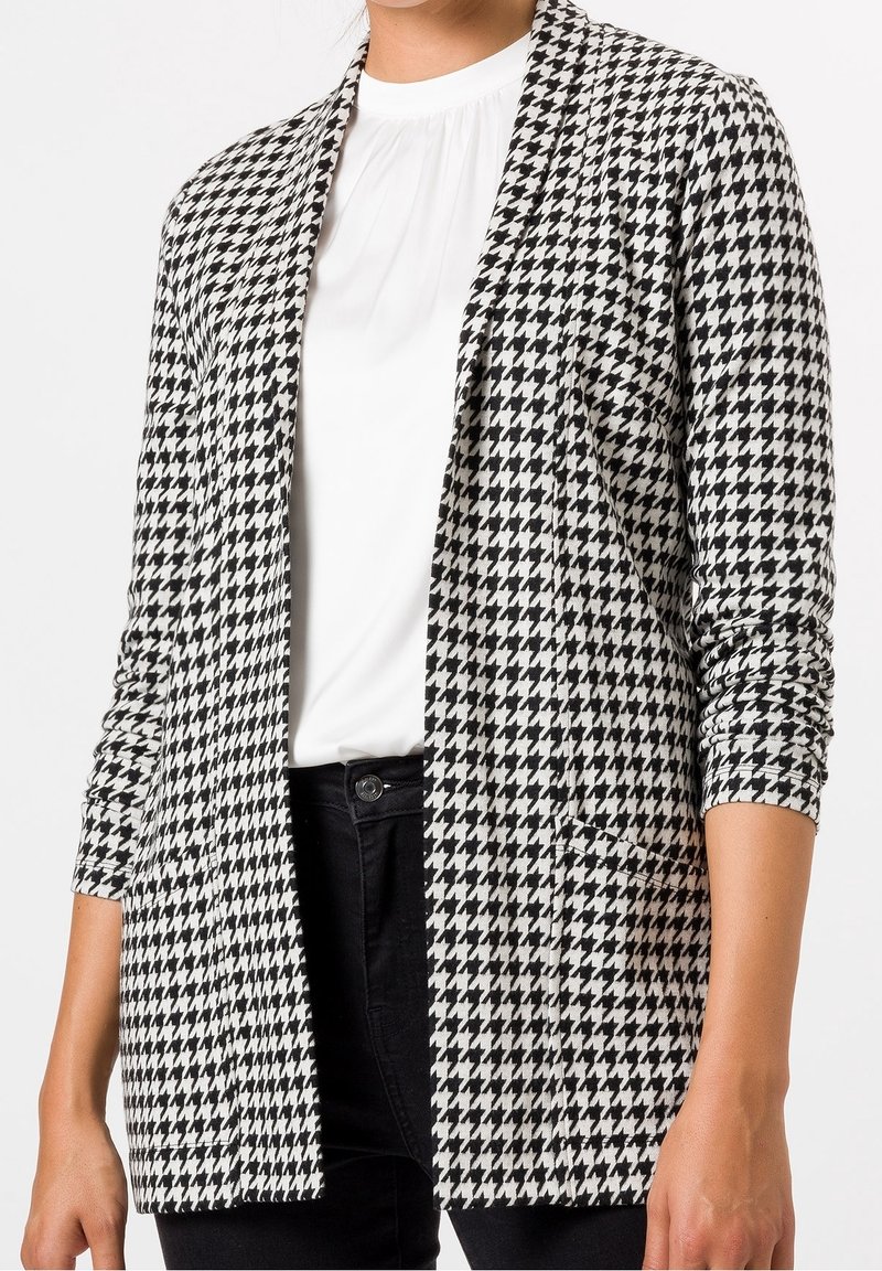 Mujer vistiendo una chaqueta de cuadro houndstooth en blanco y negro sobre una blusa blanca y pantalones negros, con los brazos relajados a los lados.