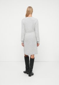 Donna che si trova di spalle, indossando un vestito a maglia grigio chiaro con maniche lunghe e stivali neri con tacco fino al ginocchio, su sfondo bianco.