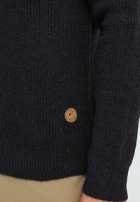 Dunkelblauer, gestrickter Pullover mit strukturiertem Finish, ausgestattet mit einem runden, hellbraunen Lederpatch mit geprägt Logo auf der unteren linken Seite.