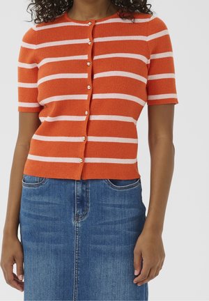 Kvinde iført en orange og hvid stribet kortærmet knapcardigan og en blå denim nederdel står mod en ensfarvet baggrund.