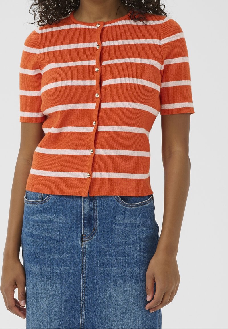 Femme portant un cardigan à manches courtes rayé orange et blanc boutonné et une jupe en jean bleue, debout devant un fond uni.