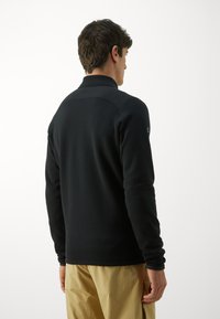 Maglia in fleece nera a maniche lunghe con colletto alto, texture liscia e design minimalista. Caratterizzata da un logo sul maniche sinistro.
