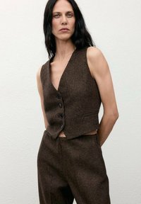Gilet di lana marrone con scollo a V profondo, dotato di quattro bottoni neri e dal design cropped. Abbinato a pantaloni marroni coordinati.