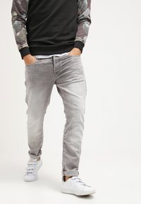 Jeans de mezclilla gris con corte slim, que presentan un lavado desgastado, cinco bolsillos y un ligero efecto de arrugado, combinados con zapatillas blancas y una sudadera negra.