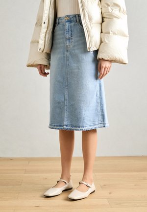 Person iført en lyseblå denim midi nederdel, beige puffjakke, beige ribbet top og cremefarvede Mary Jane sko stående på trægulv.