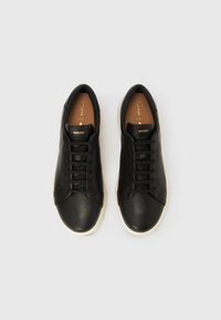 Ζευγάρι μαύρα δερμάτινα sneakers με κορδόνια και λευκές σόλες, με το εμπορικό σήμα «Geox» ορατό στις γλώσσες και τα εσωτερικά πέλματα, απεικονιζόμενα από πάνω σε λευκό φόντο.