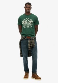 Grøn t-shirt med korte ærmer og hvid grafisk tryk, blå denim jeans og brune snørebåndsstøvler. Rutig skjorte bundet om livet.