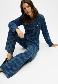 Mujer con cabello marrón ondulado sentada en el suelo, vistiendo una chaqueta de mezclilla azul con estampado y pantalones de pierna ancha a juego, con zapatos negros sin cordones.