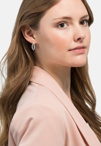 Boucle d'oreille en argent avec un design épuré et moderne. Le modèle porte un blaser rose clair. Ses cheveux sont ondulés, tombant sur une épaule.