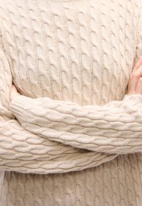 Personne portant un pull beige à torsades, bras croisés, mains reposant sur les bras opposés.