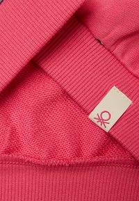 Helles pinkes Textil mit geripptem und strukturiertem Stoff. Enthält ein angenähtes Etikett mit einem pinken Logo auf einem cremefarbenen Hintergrund, das weiche Materialien präsentiert.