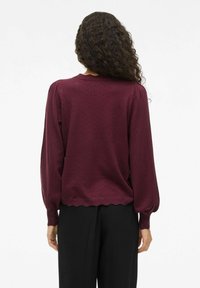 Mujer con cabello rizado vistiendo un suéter burdeos de manga larga con detalles texturizados y pantalones negros, de espaldas sobre un fondo blanco.