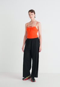 Orange bodysuit with a bow detail, paired with loose black pants. Model wearing black and red athletic shoes. Plain background.