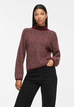 Femme portant un pull à motif chevrons bordeaux et blanc avec un pantalon noir, posant avec une main dans la poche devant un fond blanc uni.