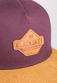 Gorra de tela color marrón con un parche de cuero color beige que presenta la inscripción "BLACKSKIES PREMIUM SUPPLIES" en relieve. La gorra tiene un visor plano y un tejido texturizado.