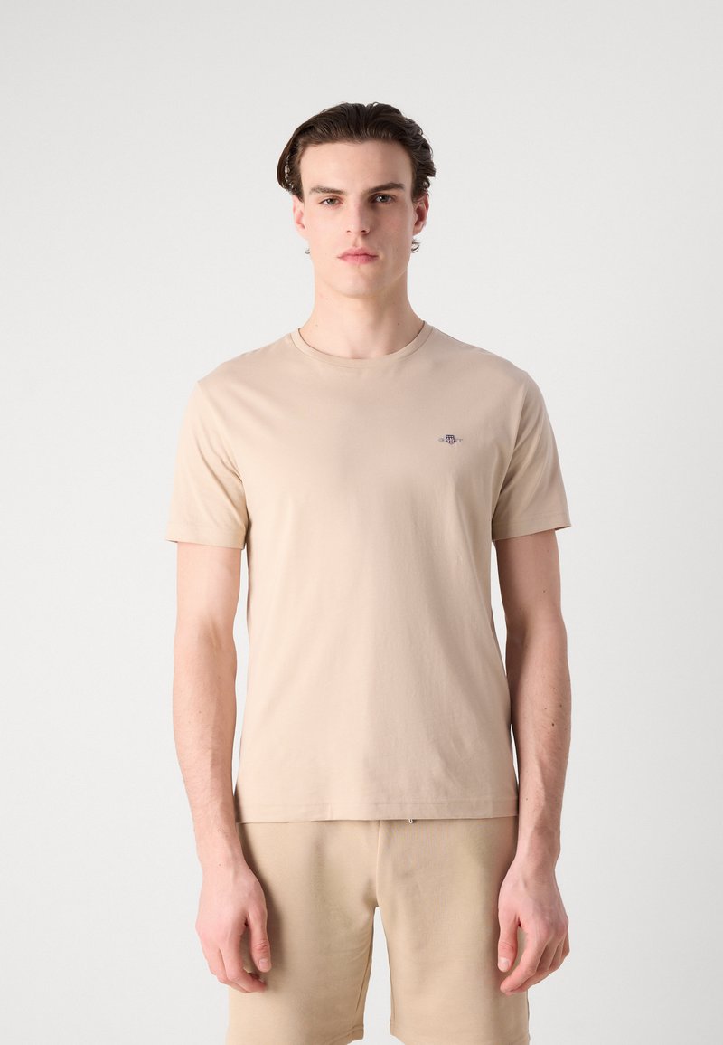 Beige T-Shirt mit kurzen Ärmeln aus leichtem Baumwollstoff, mit Rundhalsausschnitt und einem dezenten Logo-Detail auf der Brust.