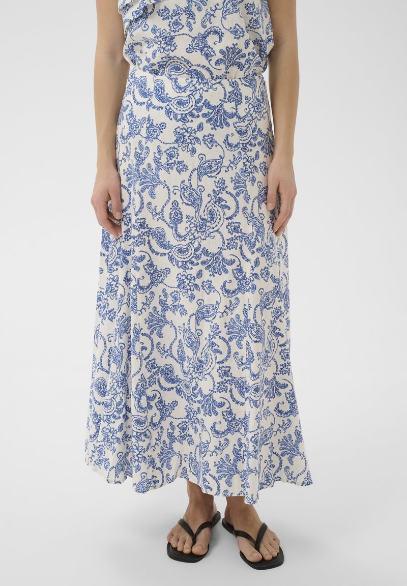 Femme portant une jupe midi à motif floral bleu et blanc associée à des sandales noires en tongs, debout devant un fond blanc.