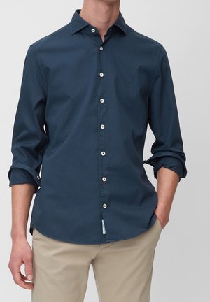 Mann trägt ein hellblaues Hemd mit hochgekrempelten Ärmeln und beigen Hosen, steht mit einer Hand in der Tasche vor einfachem Hintergrund.