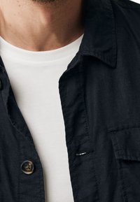 Close-up of a person wearing a white crew-neck shirt underneath an unbuttoned black collared shirt.