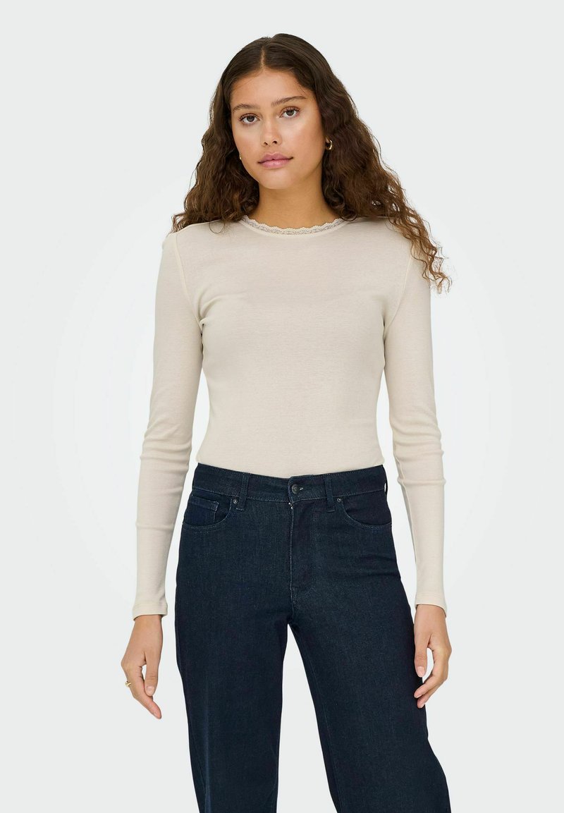 Beige langermet topp med bølget utringning, kombinert med mørkeblå denimjeans med frontlommer og klassisk passform.