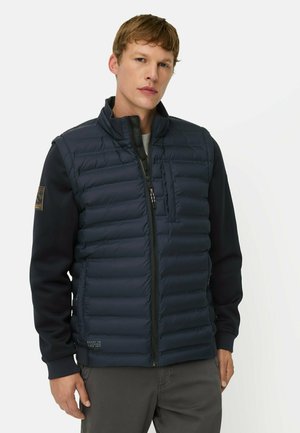 Junger Mann mit kurzen Haaren, der eine marineblaue gesteppte Weste über einem schwarzen Langarmshirt und grauen Hosen trägt, vor weißem Hintergrund stehend.