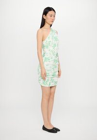 Robe à une épaule en imprimé floral vert clair sur tissu blanc, présentant des détails de ruching et une silhouette près du corps. Des ballerines noires sont portées.