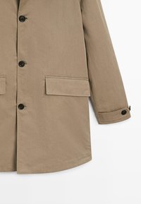 Cappotto beige con bottoni neri, tasche frontali con pattina e dettaglio a pattina con bottoni sul polsino, mostrato dalla metà del corpo fino alla fine della manica.