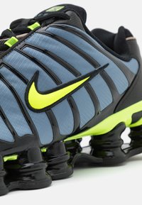 Sapatilhas de corrida Nike com um superior texturizado em cinza, detalhes em preto e um logótipo amarelo brilhante. Apresenta uma sola preta acolchoada distinta.