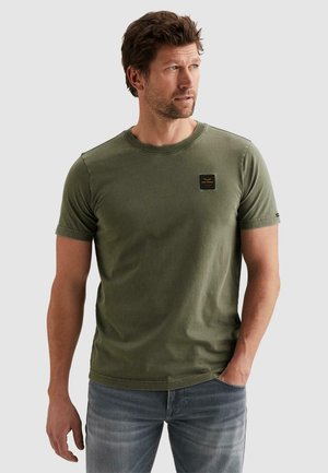 Mann trägt olivgrünes T-Shirt mit kurzen Ärmeln und ausgewaschene blaue Jeans, blickt nach rechts und hat eine Hand in der Tasche.