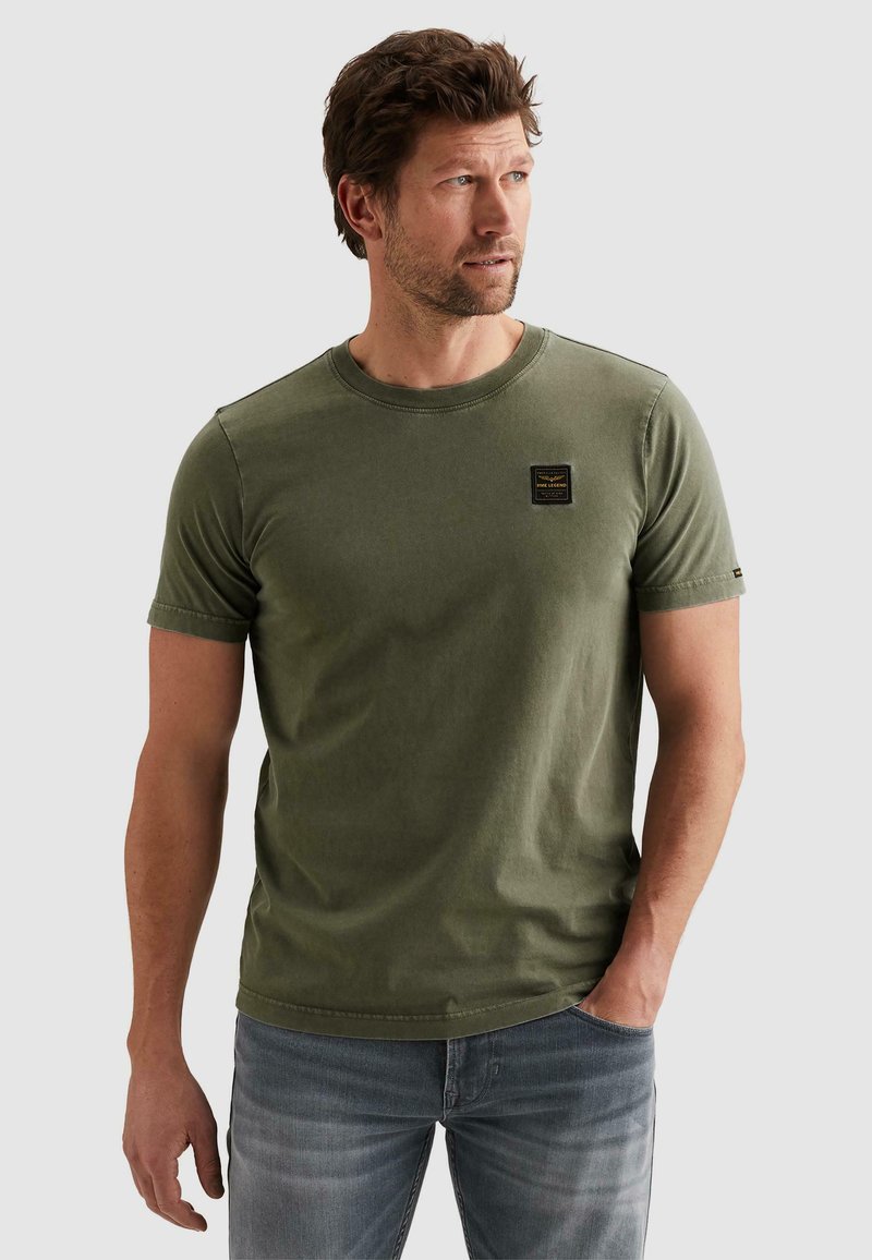 Moški v kratki olivno zeleni majici in izpranih modrih jeans hlačah, ki gleda desno, ena roka v žepu.